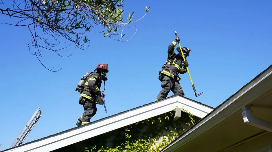 Two firefighters in full gear on a residential roof performing duties during daytime.